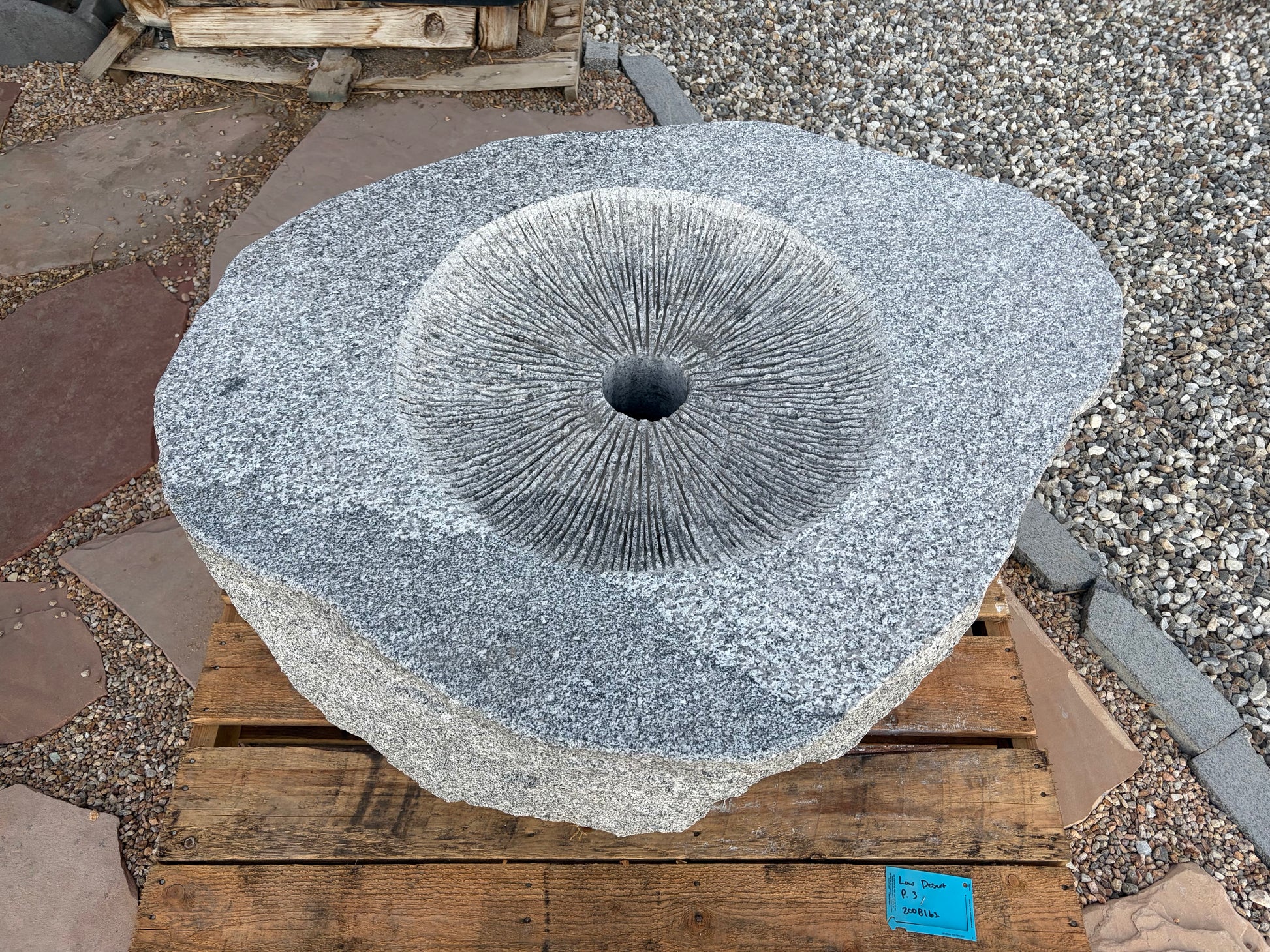 A large round gray granite stone mill wheel with a central hole and radial grooves, resting on a wooden pallet outdoors on a gravel and stone surface.