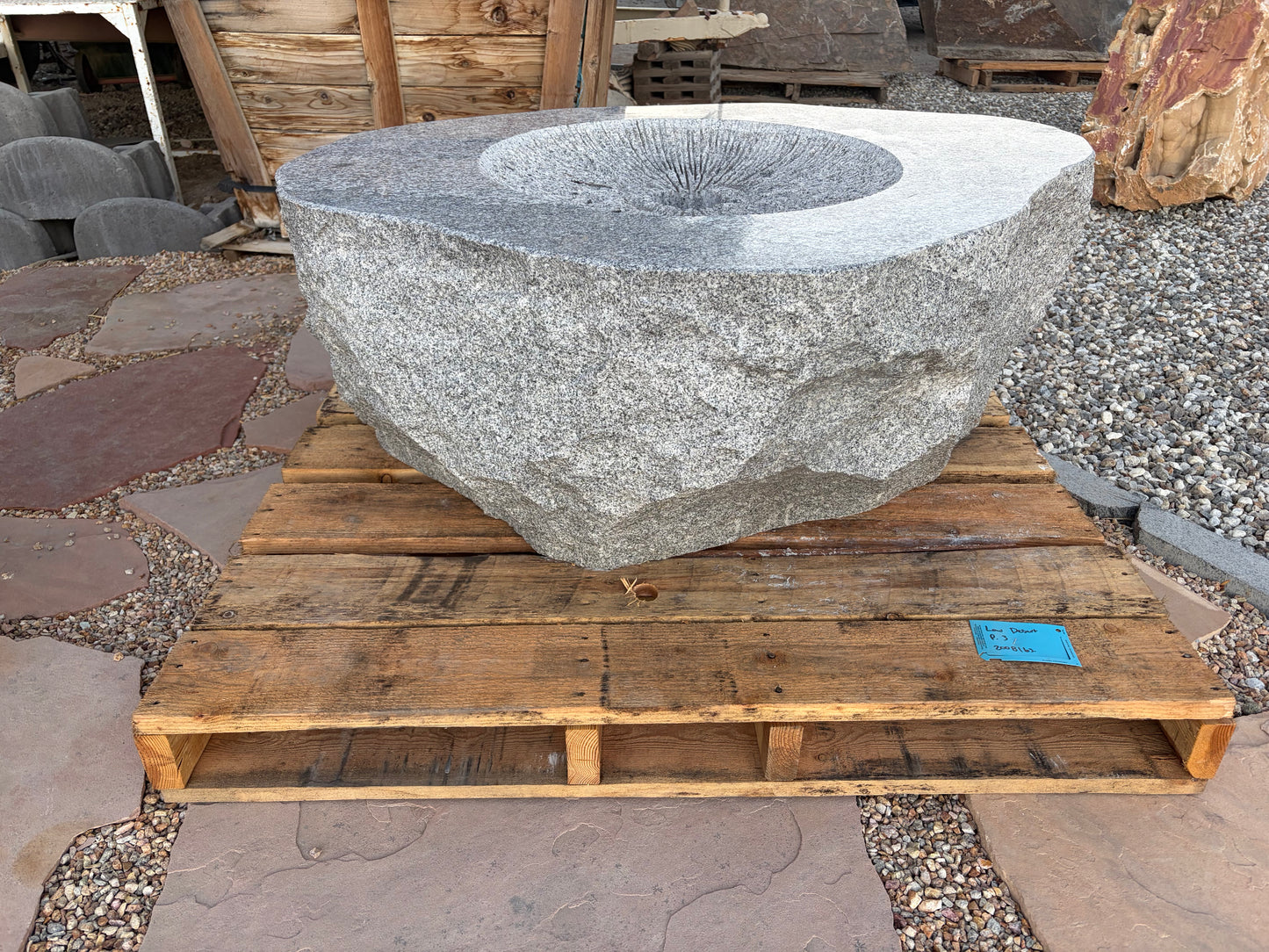 A large rough-hewn stone fountain basin made of speckled gray granite, displayed on a wooden pallet in an outdoor stone yard.